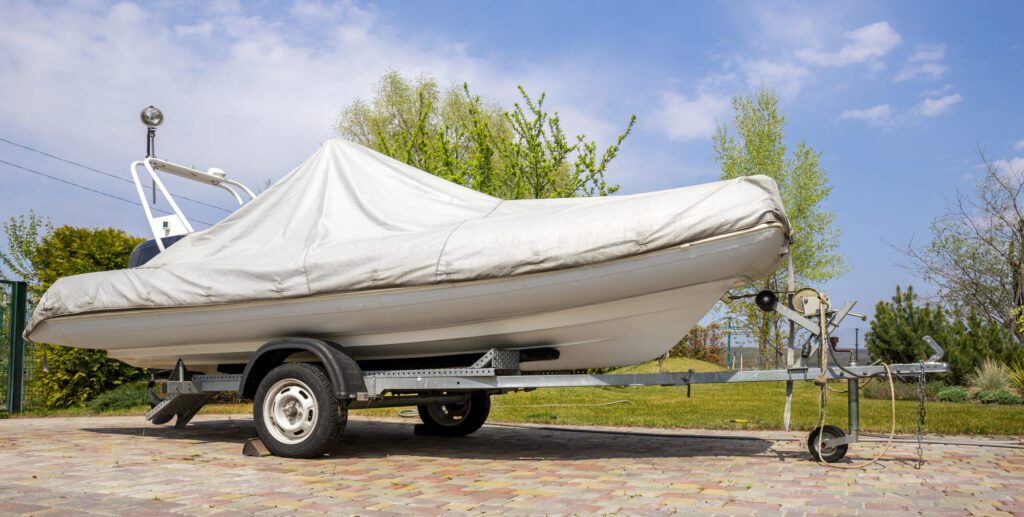 boat covered with grey or white protection tarp standing on steel semi trailer at home backyard on bright sunny day with blue sky on background. Boat vessel storage.